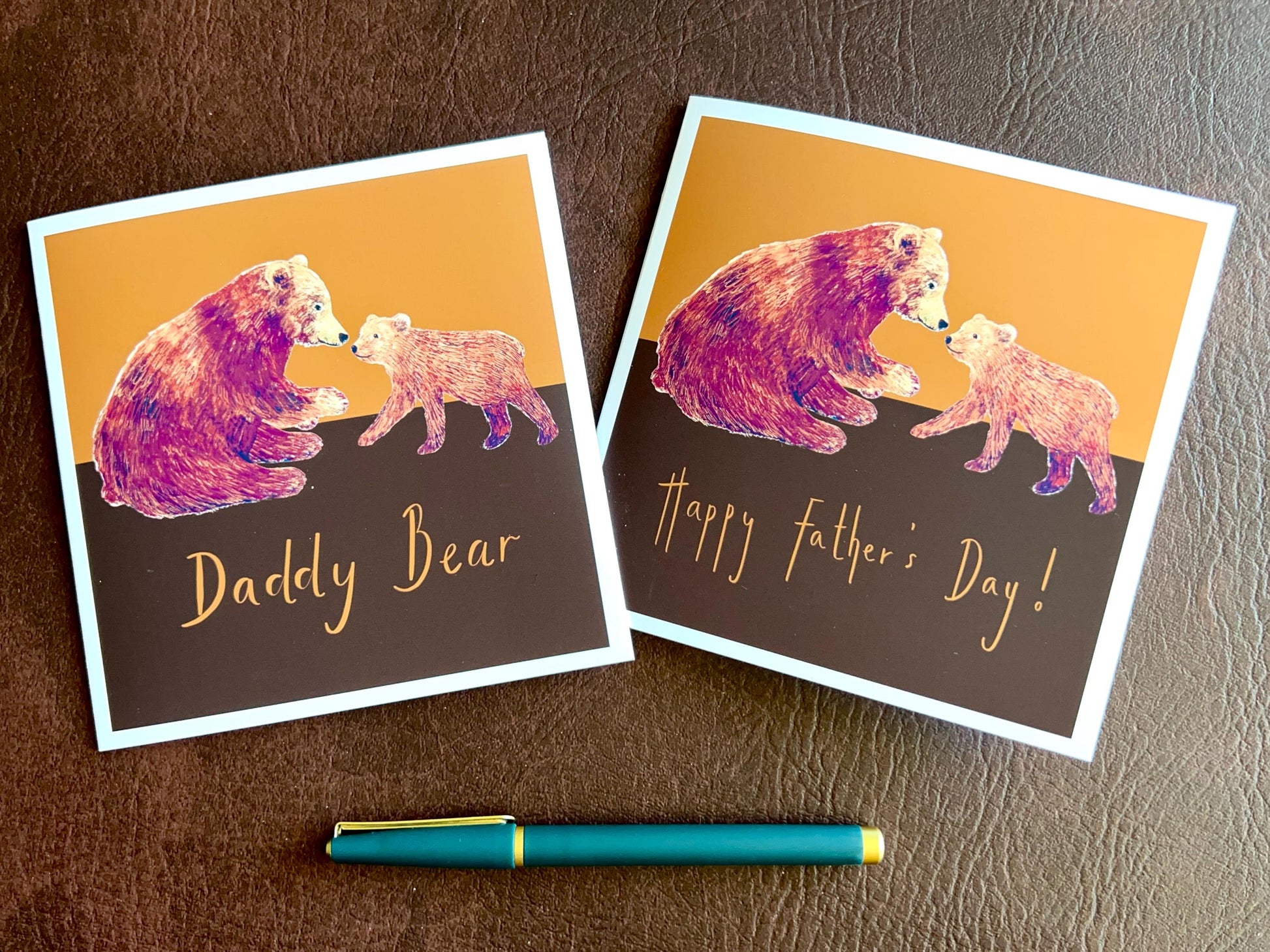 Flat lay image showing two artisan Fathers Day card designs featuring a brown bear and bear cub on a warm brown and burnt orange background. One card reads 'Happy Father’s Day' and the other reads 'Daddy Bear', displayed side by side in a styled composition.