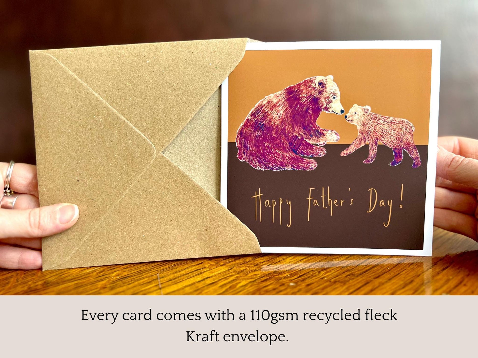 Infographic showing an artisan Fathers Day card featuring a brown bear and bear cub on a warm brown and burnt orange background being removed from a kraft envelope, with text explaining that every card comes with a 110gsm recycled fleck kraft envelope.