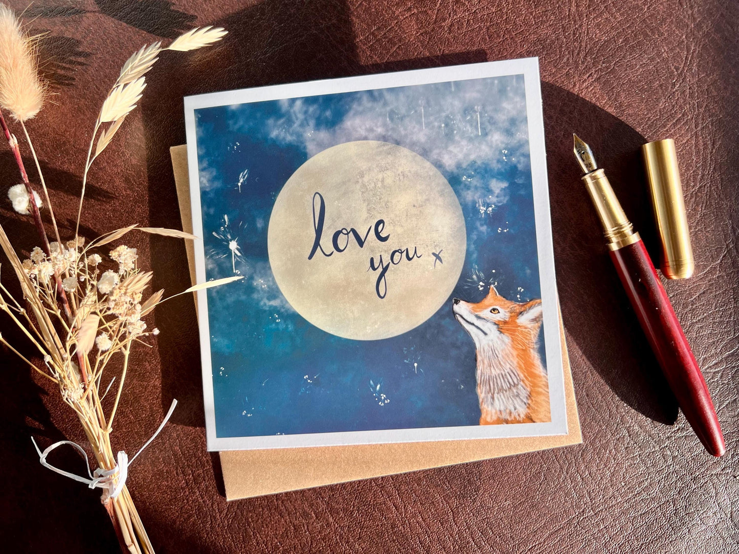 Flat lay image of an artisan romantic card featuring a red fox looking up at a glowing full moon in a blue starry night sky with handwritten style text reading 'love you x', shown laid flat on a tabletop next to a pen and dried flowers in a styled composition.