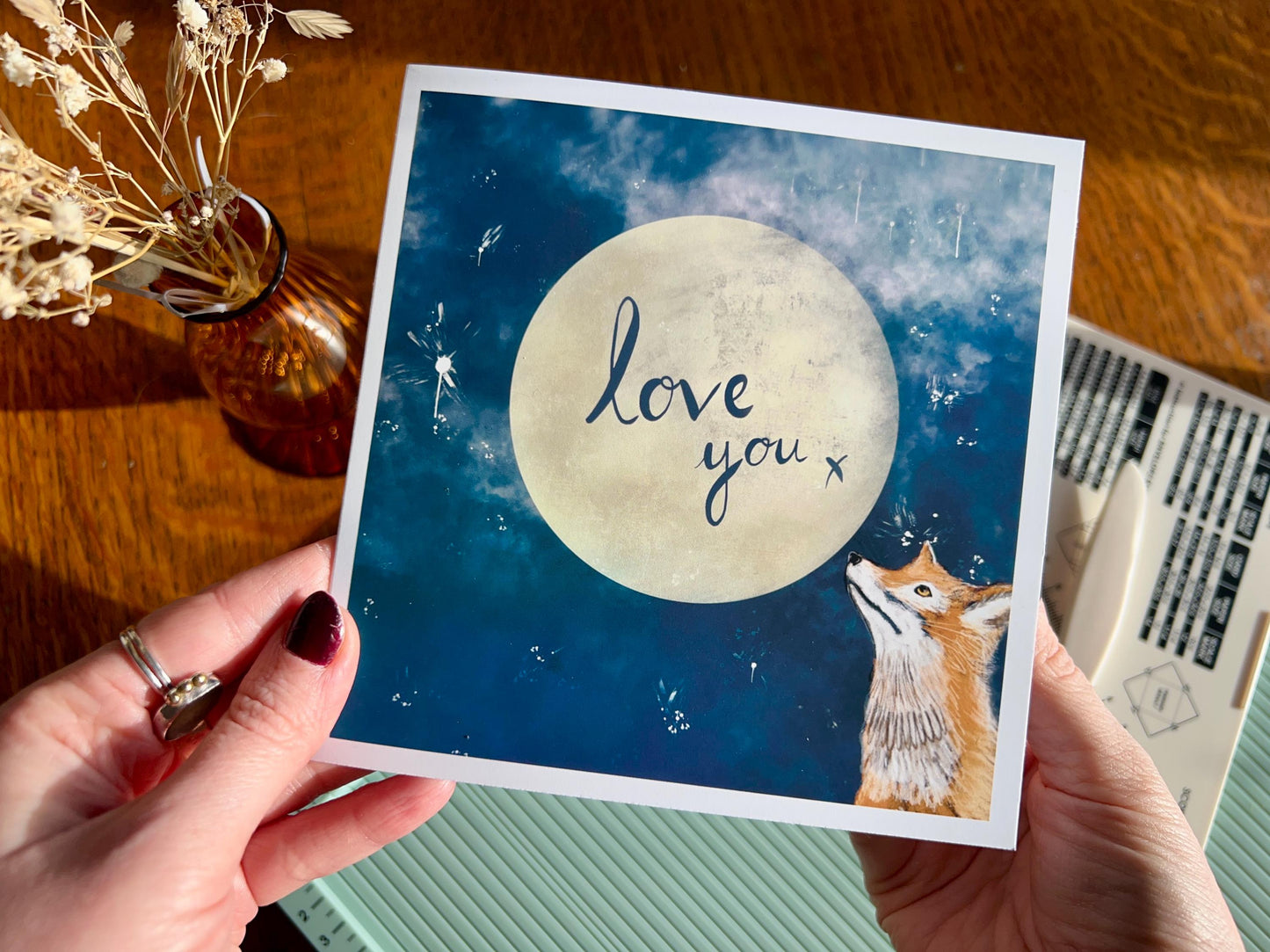 Hands holding an artisan romantic card above a scoring board, showing the handcrafted production process. The design features a red fox looking up at a glowing full moon in a blue starry night sky with handwritten style text reading 'love you x' inside the moon.
