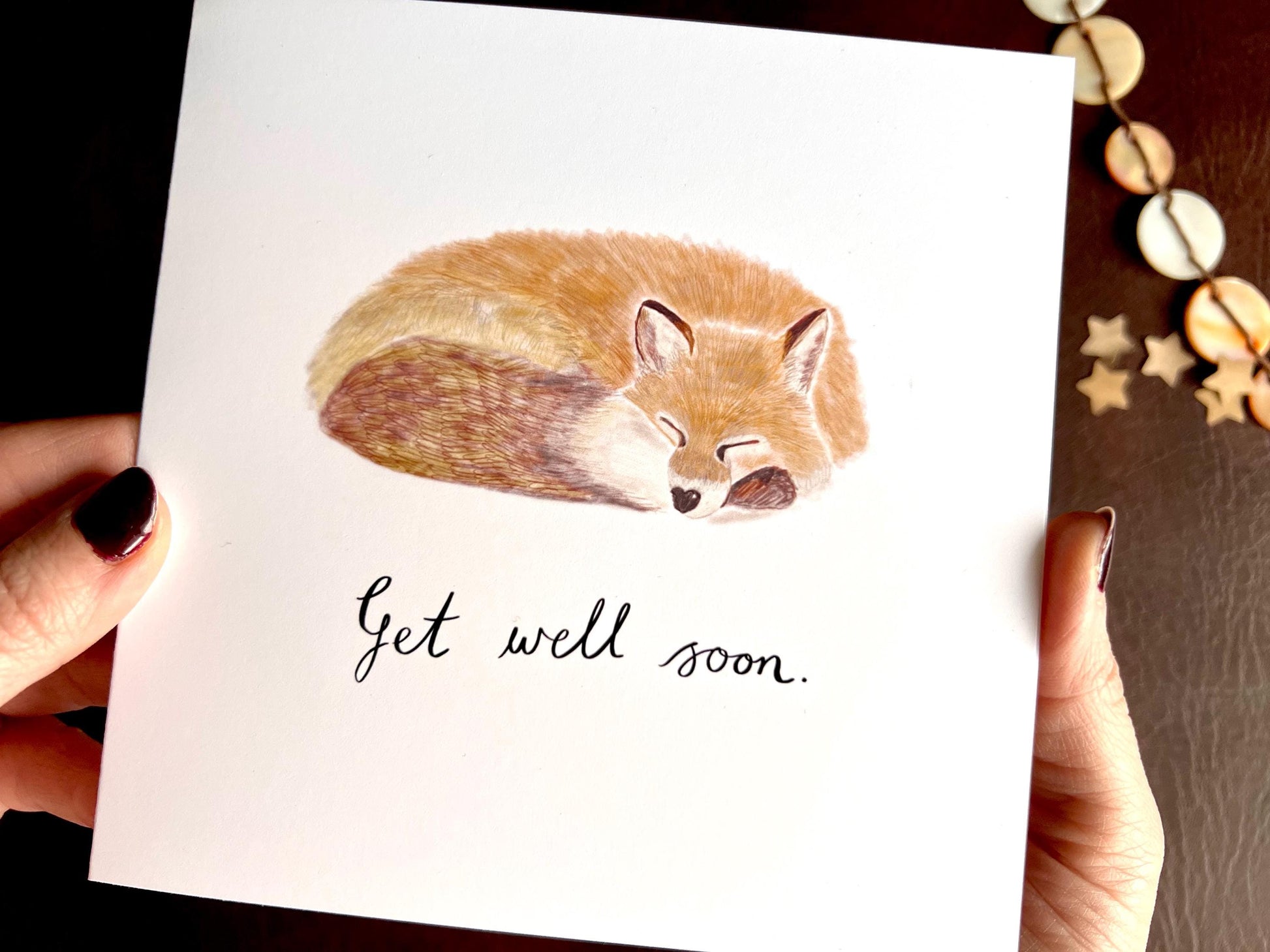 Close up image of hands holding an artisan get well soon card being removed from a kraft envelope, featuring a sleeping fox illustration on a plain white background with handwritten style text reading 'Get well soon'.