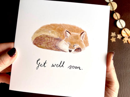 Close up image of hands holding an artisan get well soon card being removed from a kraft envelope, featuring a sleeping fox illustration on a plain white background with handwritten style text reading 'Get well soon'.