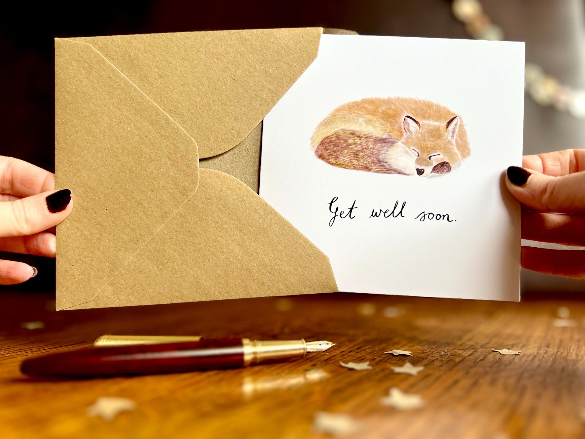 Front view image of hands holding an artisan get well soon card being removed from a kraft envelope, featuring a sleeping fox illustration on a plain white background with handwritten style 'Get well soon' text.