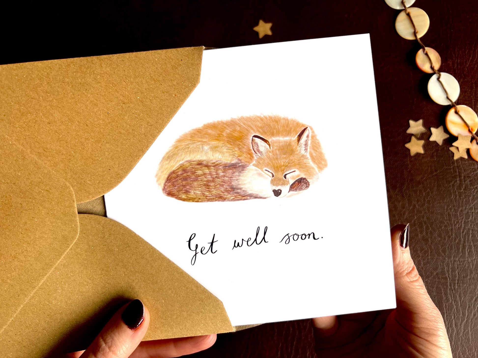 Hands holding an artisan get well soon card being removed from a kraft envelope, featuring an illustration of a sleeping fox on a plain white background, designed as a calming and comforting sympathy style card.