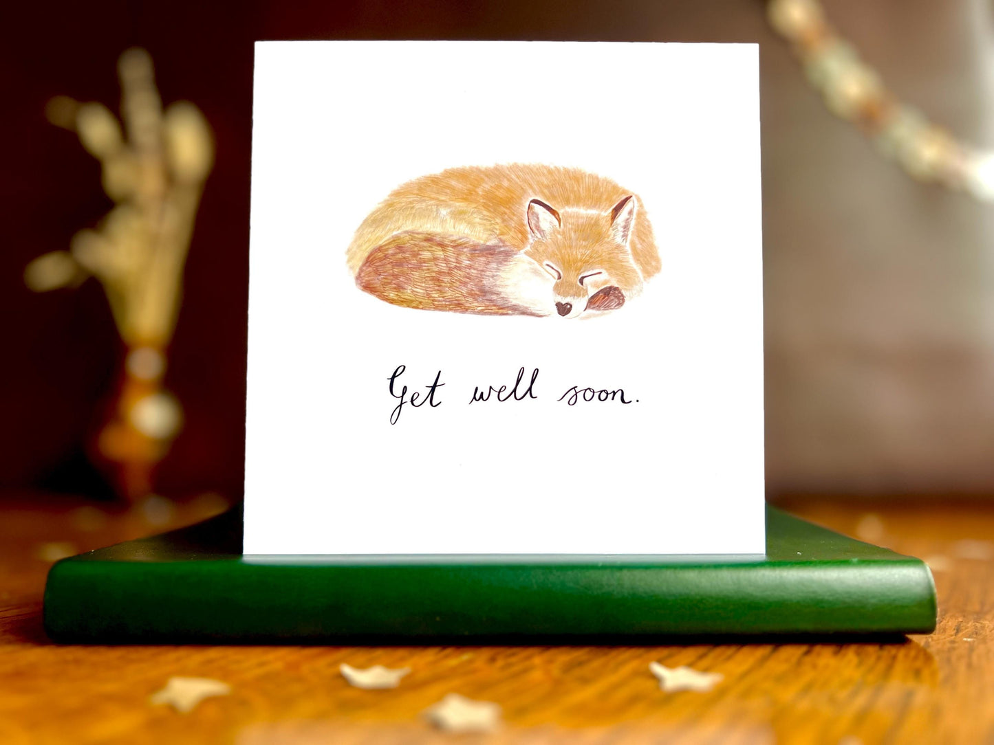 Artisan get well soon card featuring a sleeping fox illustration on a plain white background with handwritten style text reading 'Get well soon' underneath, shown standing upright on a green leatherette book in a styled lifestyle setting.