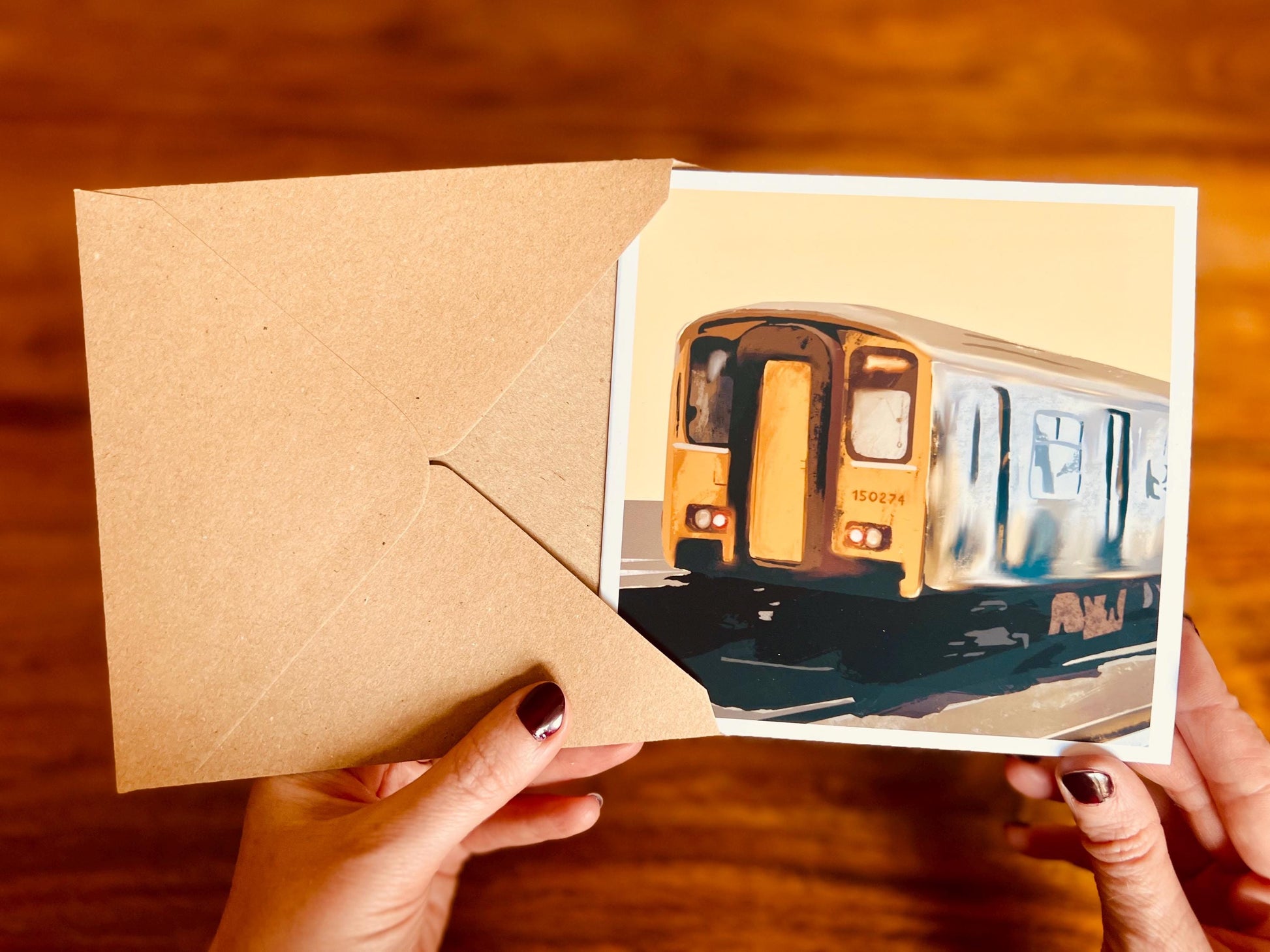Aerial view of hands holding an artisan greeting card being removed from a high quality kraft envelope, showing the front design of the card as it is being viewed and handled.