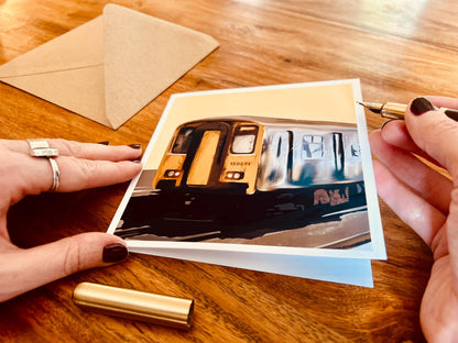Hand opening an artisan greeting card with a fountain pen ready to write inside, showing the card partially open so the front design is still visible. A kraft envelope is laid flat on the tabletop in the background.