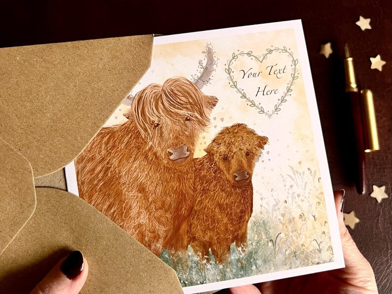 Hands holding an artisan greeting card being removed from a kraft envelope and lifted towards the camera, featuring two Highland cows on a lemon yellow background. The floral heart contains the text 'Your Text Here' to demonstrate customisation.