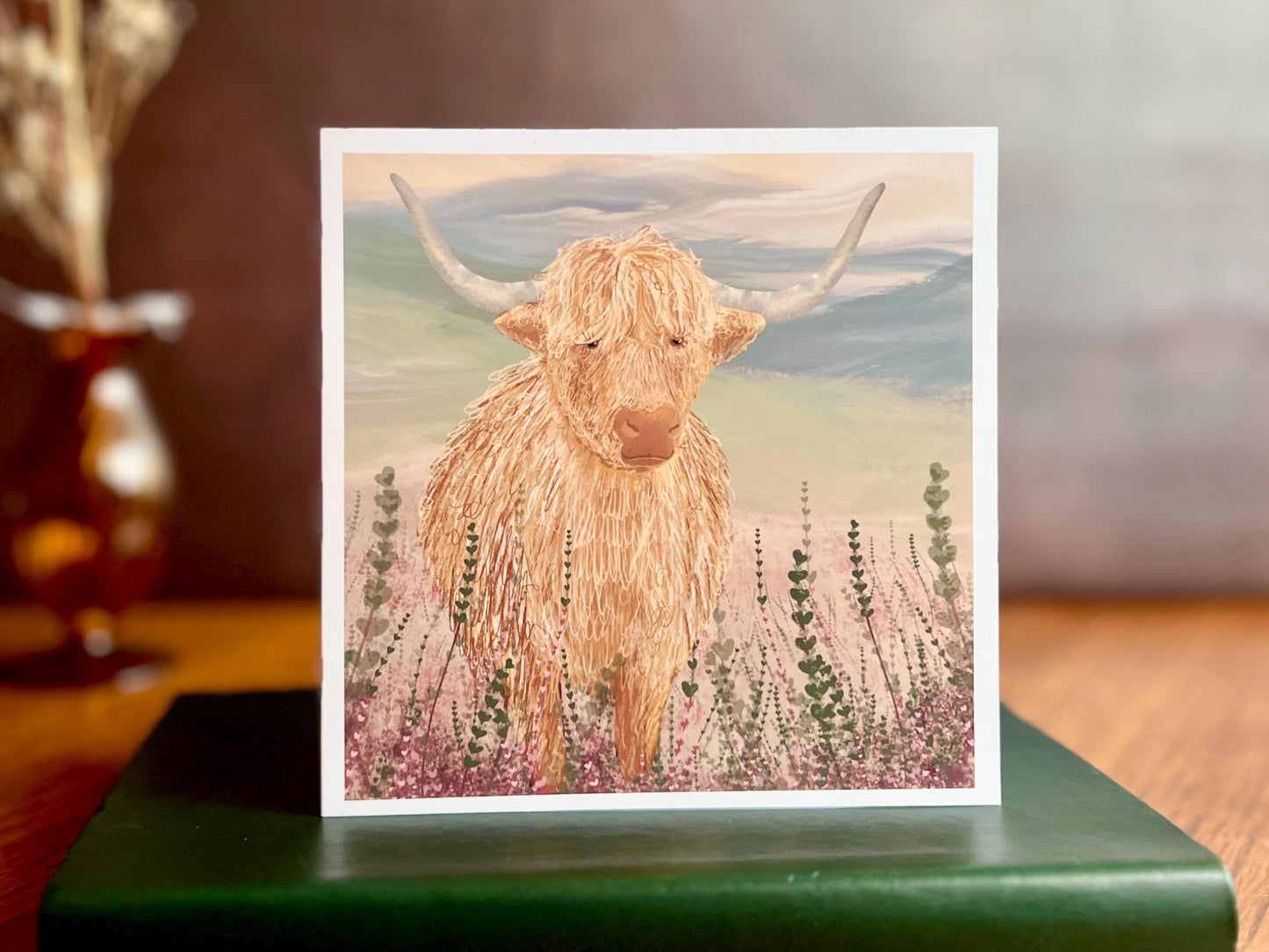 Artisan greeting card featuring a single highland cow standing among purple heather with a sage green moorland background and no text, shown standing upright on a green leatherette book in a styled lifestyle display.