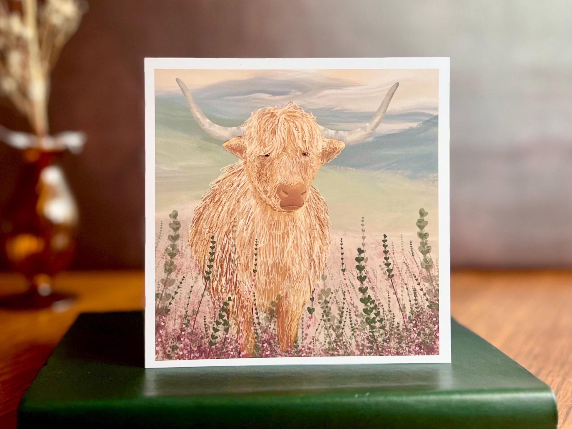 Artisan greeting card featuring a single highland cow standing among purple heather with a sage green moorland background and no text, shown standing upright on a green leatherette book in a styled lifestyle display.