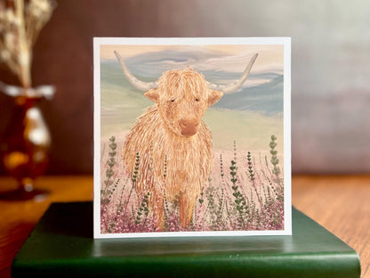 Artisan greeting card featuring a single highland cow standing among purple heather with a sage green moorland background and no text, shown standing upright on a green leatherette book in a styled lifestyle display.