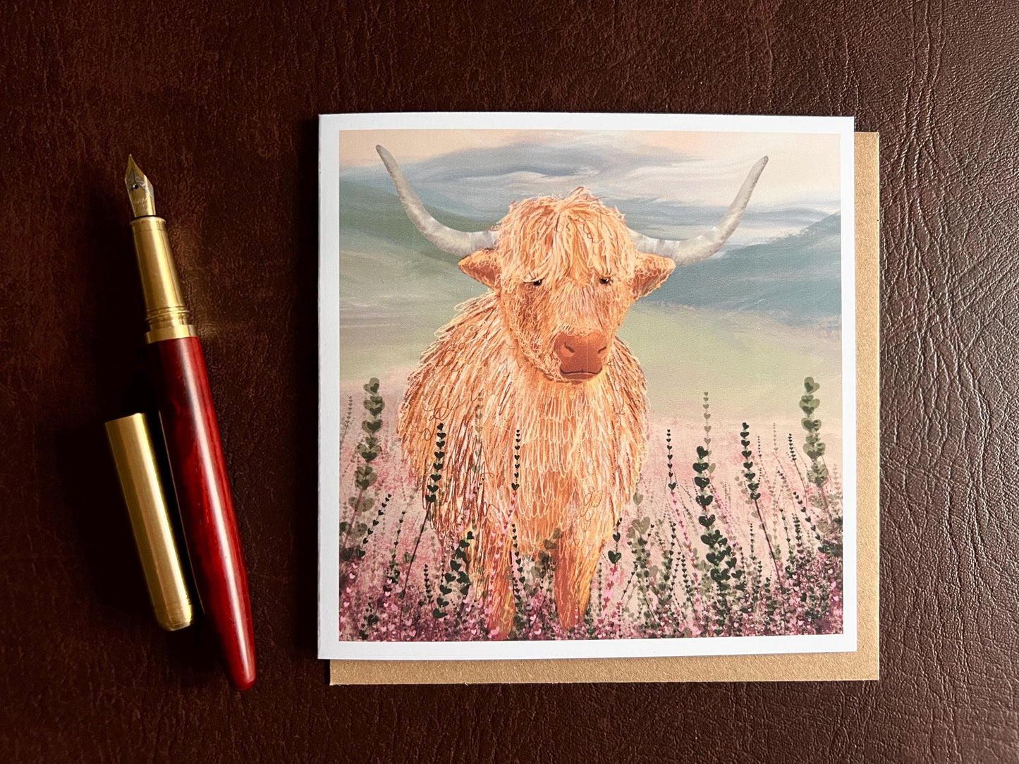 Flat lay image of an artisan greeting card featuring a single highland cow standing among purple heather with a sage green moorland background and no text, shown laid flat on a tabletop in a styled composition.