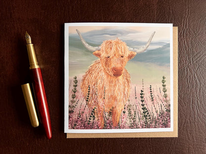 Flat lay image of an artisan greeting card featuring a single highland cow standing among purple heather with a sage green moorland background and no text, shown laid flat on a tabletop in a styled composition.
