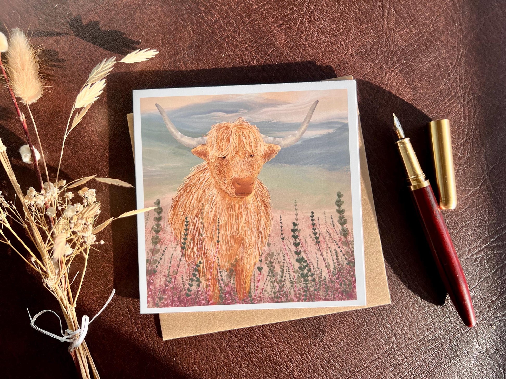 Flat lay image of an artisan greeting card featuring a single highland cow standing among purple heather with a sage green moorland background and no text, shown laid flat on a tabletop with a pen and dried flowers in a styled composition.