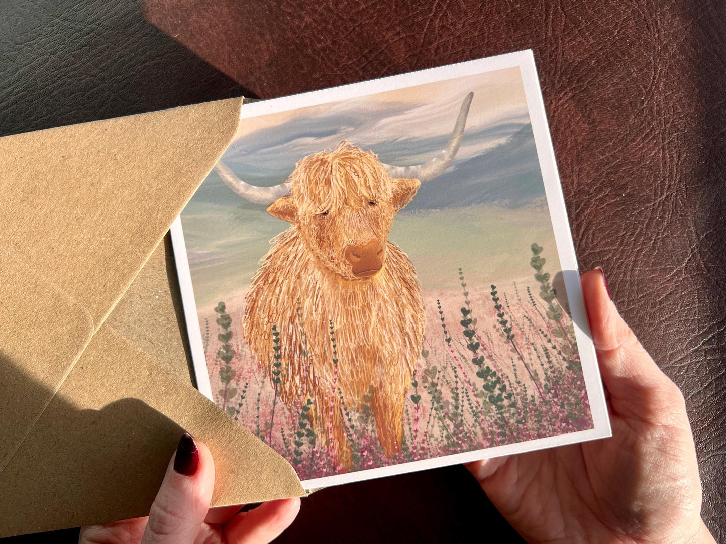 Hands holding an artisan greeting card being removed from a kraft envelope, featuring a single highland cow illustration standing among purple heather with a sage green moorland background. The design has no text on the front and focuses on a minimalist animal illustration style.