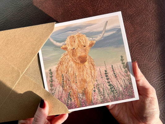 Hands holding an artisan greeting card being removed from a kraft envelope, featuring a single highland cow illustration standing among purple heather with a sage green moorland background. The design has no text on the front and focuses on a minimalist animal illustration style.