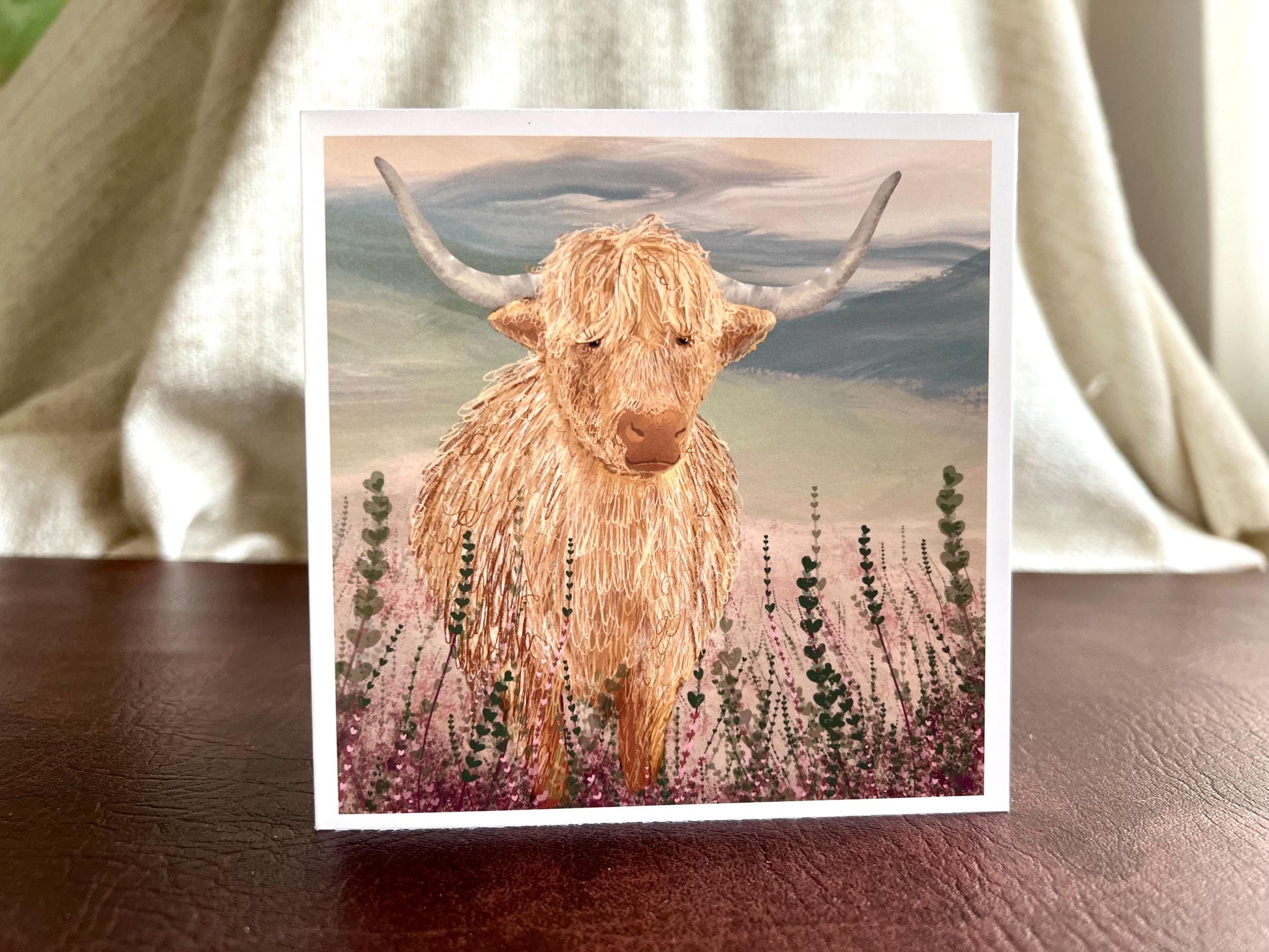 Artisan greeting card featuring a single highland cow standing among purple heather with a sage green moorland background and no text, shown standing upright on a leatherette tabletop in a styled lifestyle display.