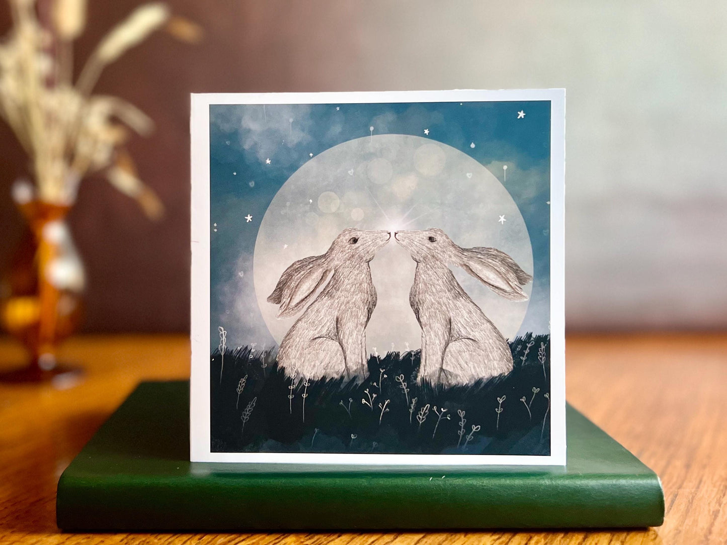 Artisan art card featuring two white hares nose to nose in front of a glowing full moon beneath a starry night sky, shown standing upright on a green leatherette book in a styled lifestyle display.