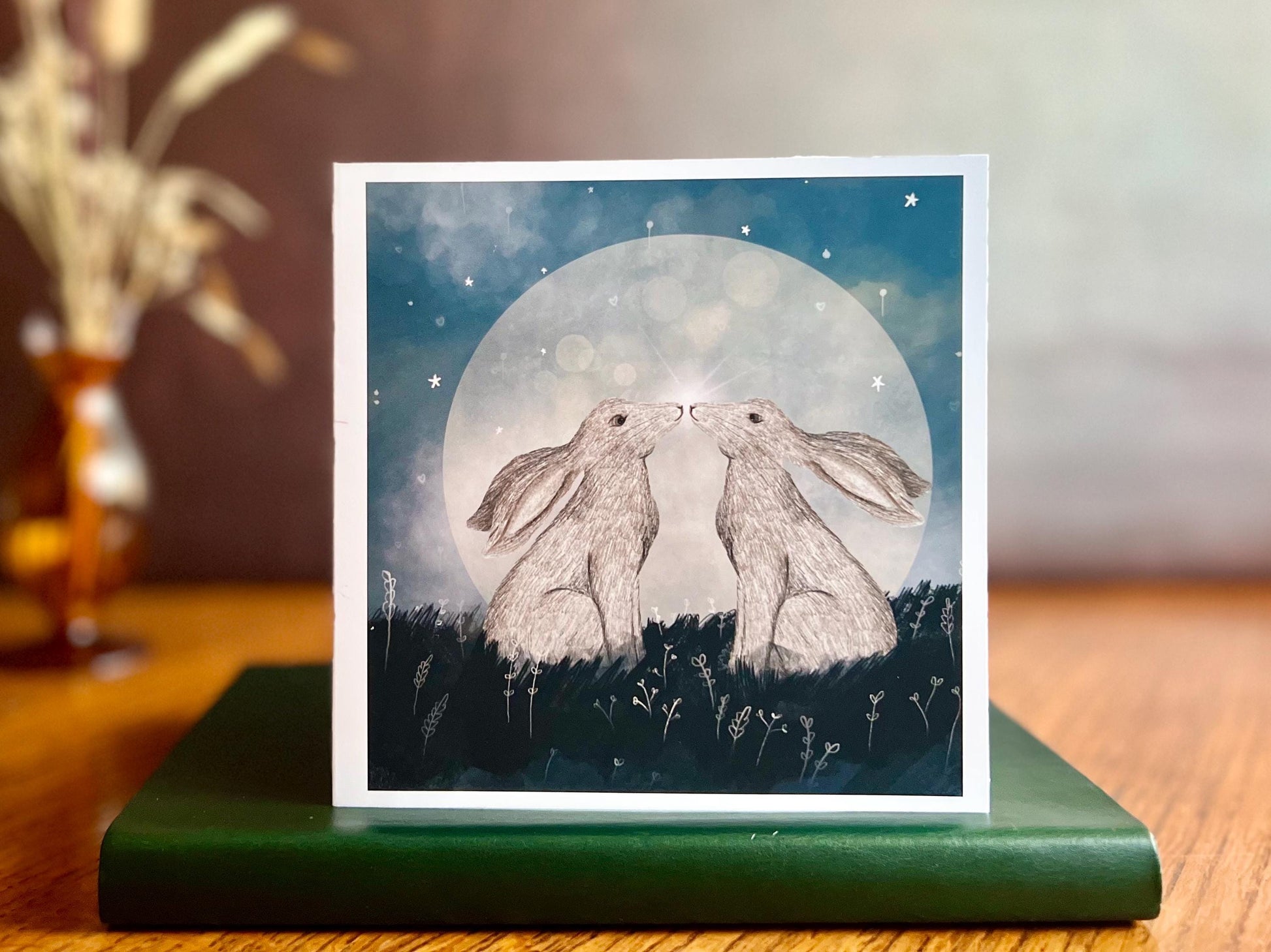 Artisan art card featuring two white hares nose to nose in front of a glowing full moon beneath a starry night sky, shown standing upright on a green leatherette book in a styled lifestyle display.
