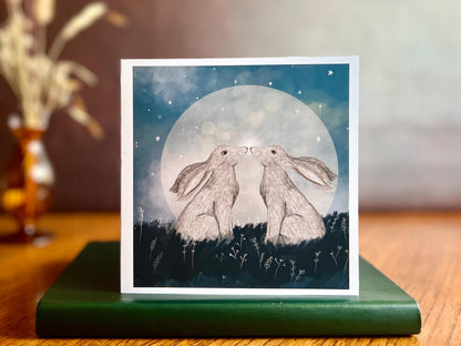 Artisan art card featuring two white hares nose to nose in front of a glowing full moon beneath a starry night sky, shown standing upright on a green leatherette book in a styled lifestyle display.