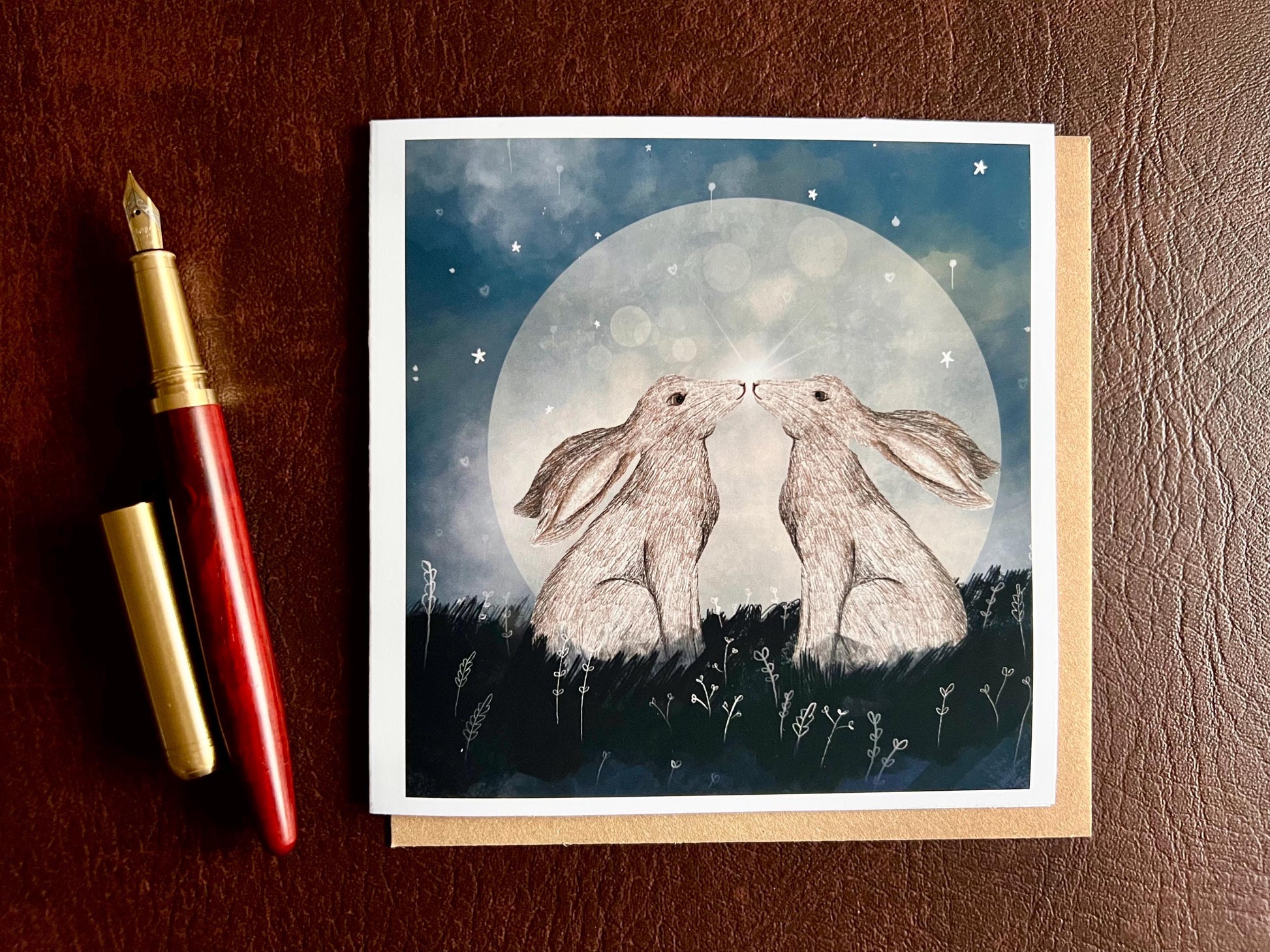 Flat lay image of an artisan art card featuring two white hares nose to nose in front of a glowing full moon beneath a starry night sky, shown laid flat on a tabletop next to a pen in a styled composition.