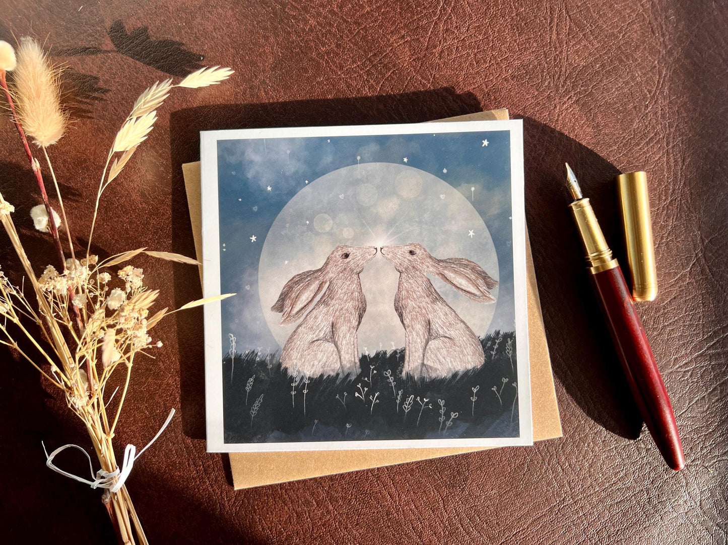 Flat lay image of an artisan art card featuring two white hares nose to nose in front of a glowing full moon beneath a starry night sky, shown laid flat on a tabletop with a pen and dried flowers in a styled composition.