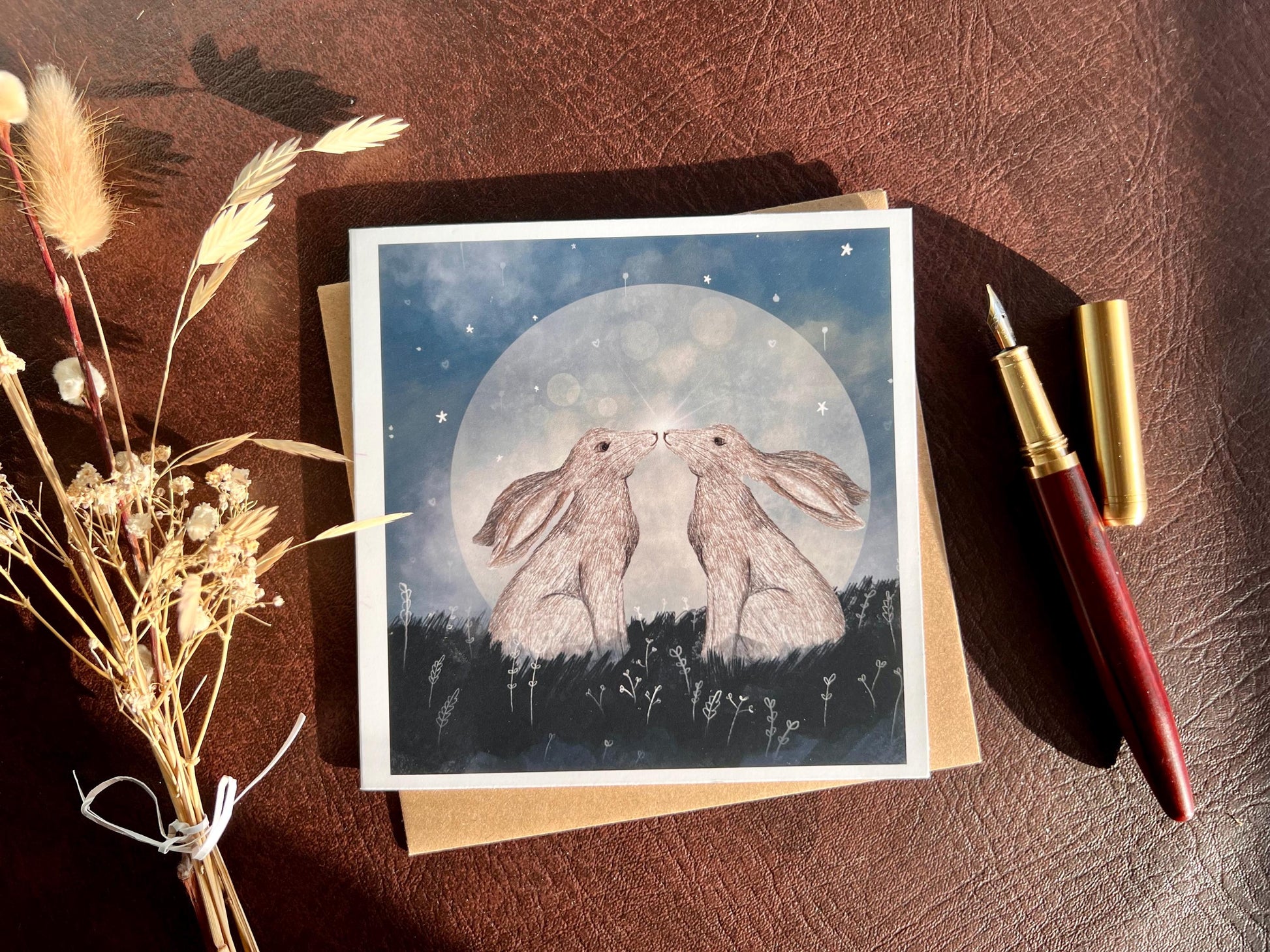 Flat lay image of an artisan art card featuring two white hares nose to nose in front of a glowing full moon beneath a starry night sky, shown laid flat on a tabletop with a pen and dried flowers in a styled composition.