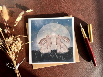Flat lay image of an artisan art card featuring two white hares nose to nose in front of a glowing full moon beneath a starry night sky, shown laid flat on a tabletop with a pen and dried flowers in a styled composition.