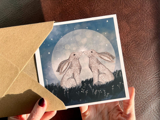 Hands holding an artisan art card being removed from a kraft envelope, featuring two white hares nose to nose in front of a glowing full moon beneath a starry night sky. The design has a mystical romantic feel representing love and companionship.
