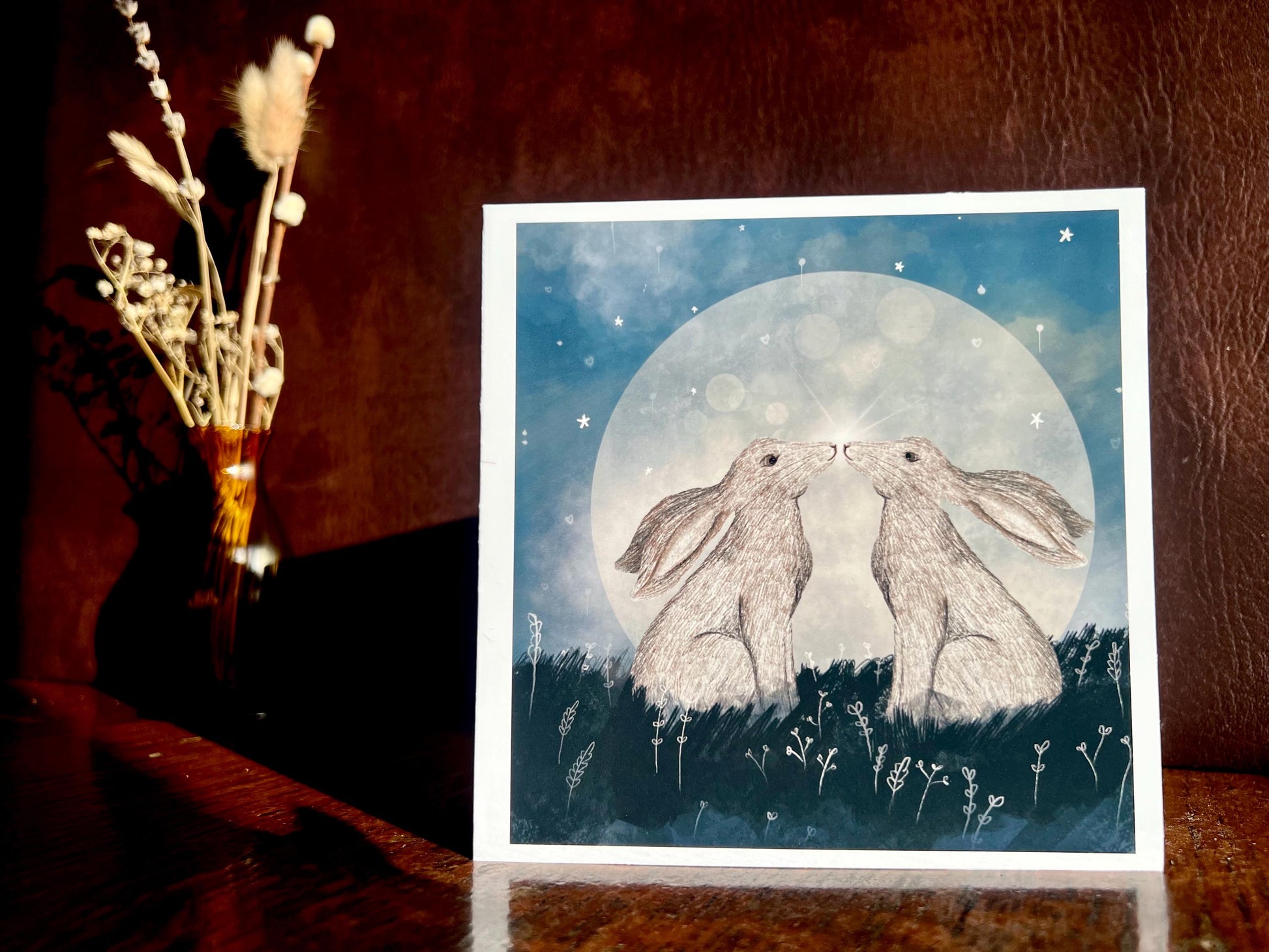 Artisan art card featuring two white hares nose to nose in front of a glowing full moon beneath a starry night sky, shown standing upright on a tabletop in a styled lifestyle display.