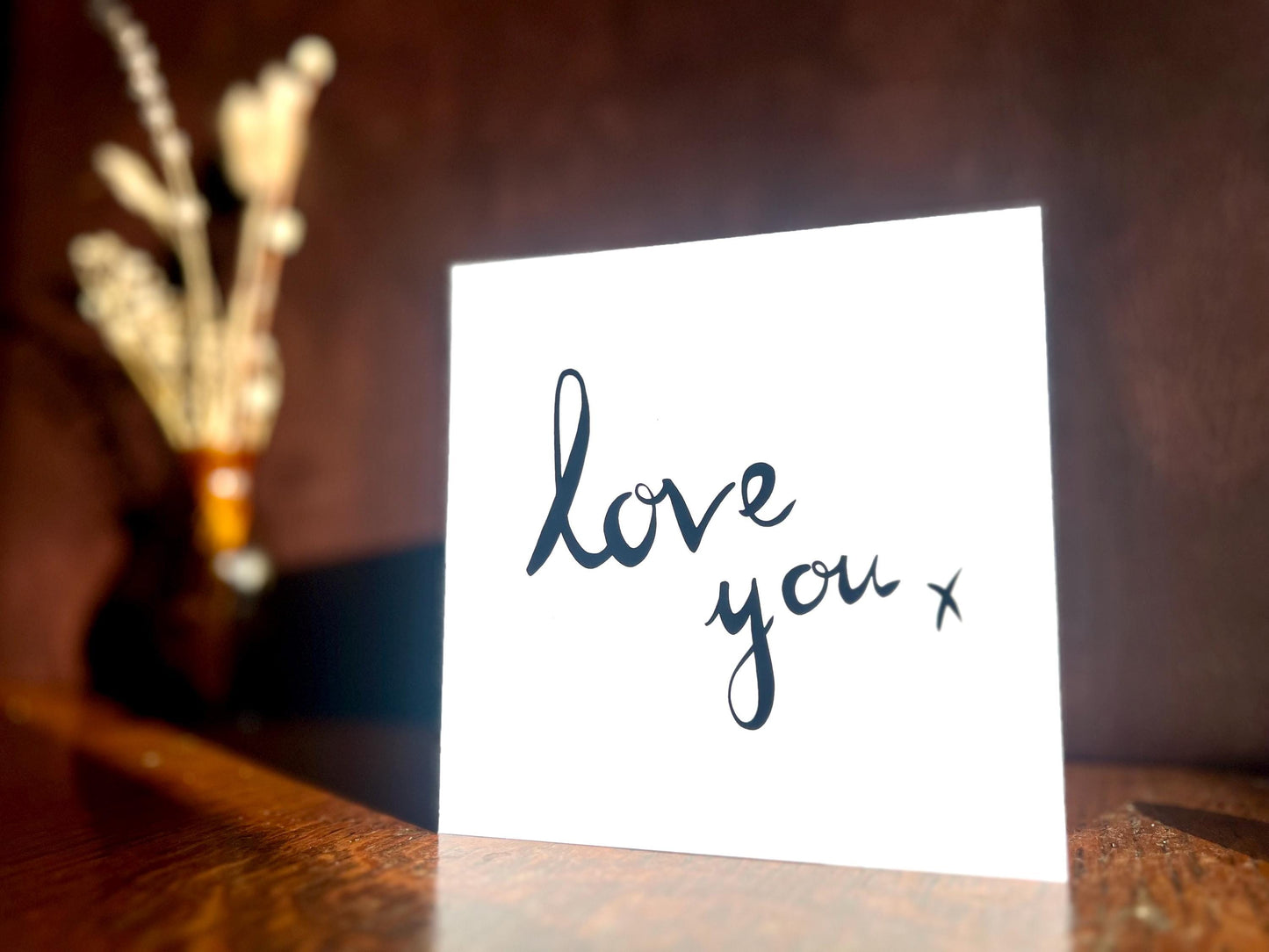 Artisan monochrome love card featuring a clean white background with the words 'love you x' in black cursive handwritten style font, shown standing upright on a tabletop in a simple styled display.