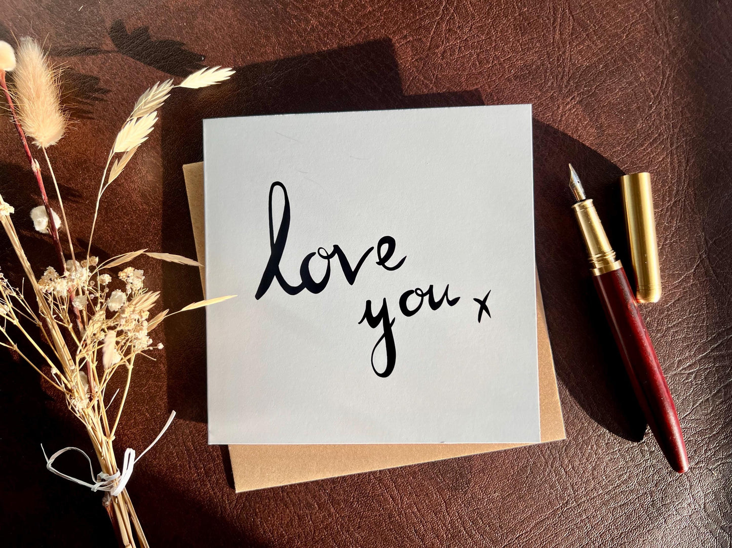 Flat lay image of an artisan monochrome love card featuring a clean white background with the words 'love you x' in black cursive handwritten style font, shown laid flat on a tabletop with a pen and dried flowers in a styled composition.