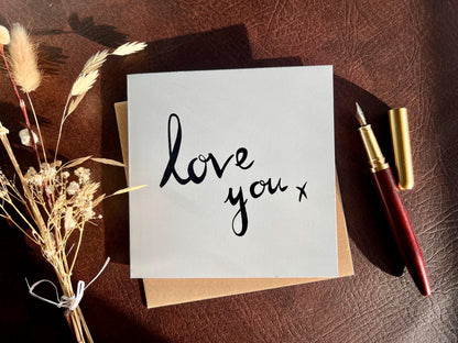 Flat lay image of an artisan monochrome love card featuring a clean white background with the words 'love you x' in black cursive handwritten style font, shown laid flat on a tabletop with a pen and dried flowers in a styled composition.