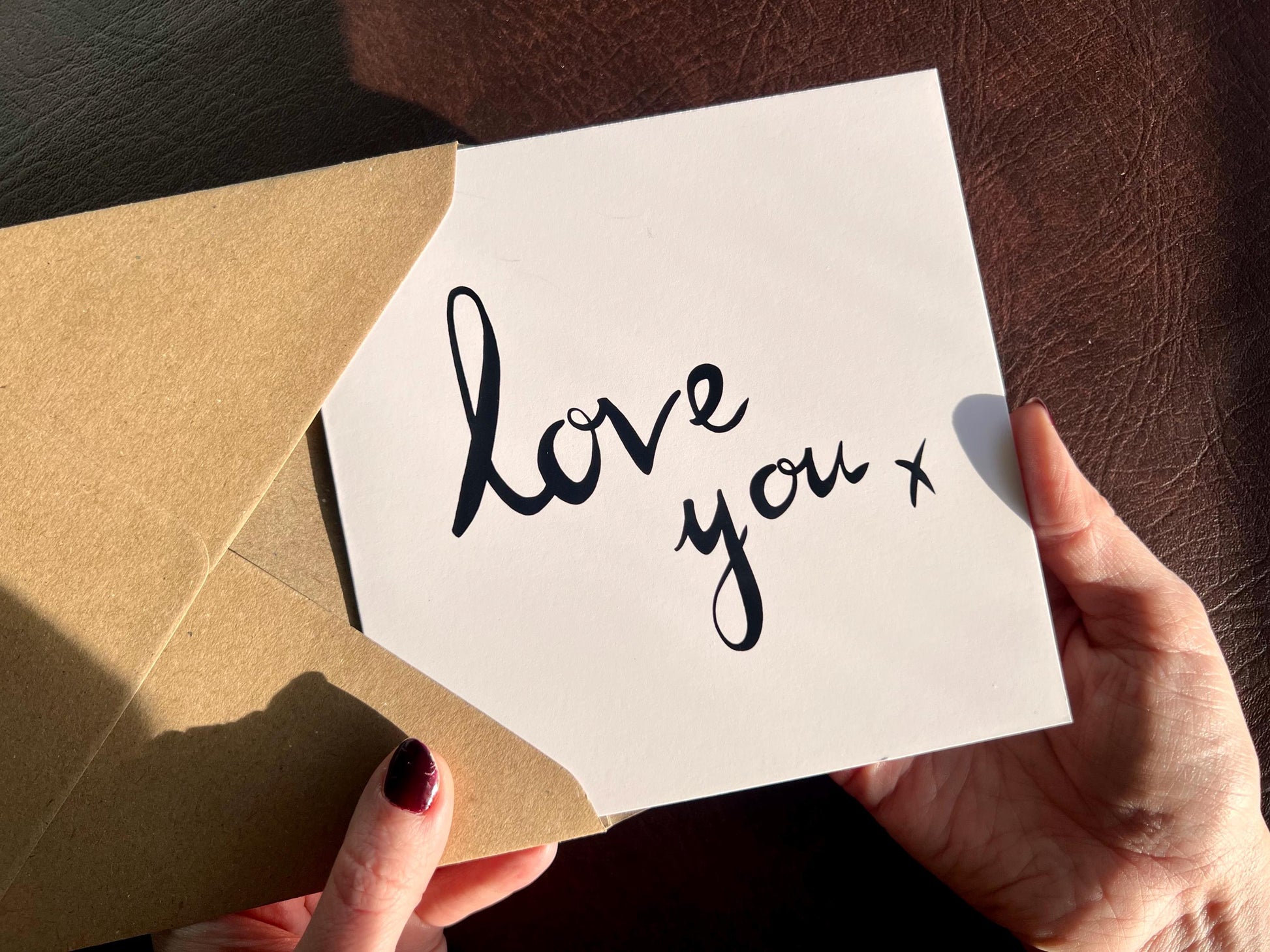 Hands holding an artisan monochrome love card being removed from a kraft envelope, featuring a clean white background with the words 'love you x' written in a black cursive handwritten style font, designed as a simple romantic card for a partner.