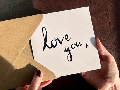 Hands holding an artisan monochrome love card being removed from a kraft envelope, featuring a clean white background with the words 'love you x' written in a black cursive handwritten style font, designed as a simple romantic card for a partner.
