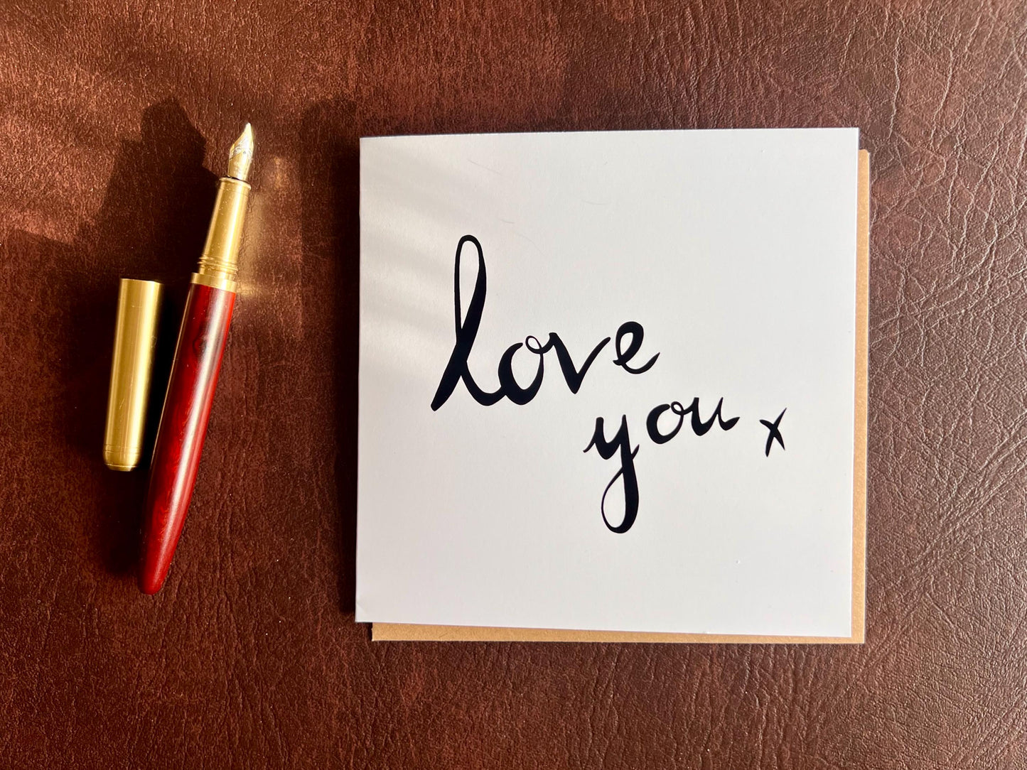 Flat lay image of an artisan monochrome love card featuring a clean white background with the words 'love you x' in black cursive handwritten style font, shown laid flat on a leatherette surface next to a pen in a styled composition.