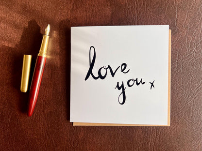 Flat lay image of an artisan monochrome love card featuring a clean white background with the words 'love you x' in black cursive handwritten style font, shown laid flat on a leatherette surface next to a pen in a styled composition.
