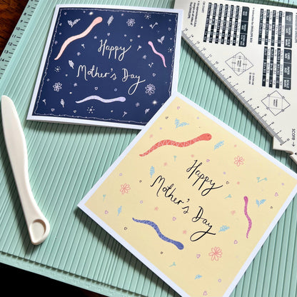 Flat lay image showing two artisan Mother’s Day card designs featuring playful cartoon snakes with handwritten style 'Happy Mother’s Day' text. One version is on a dark navy blue background and the other on a lemon yellow background, both displayed on a scoring board to highlight their handcrafted nature.