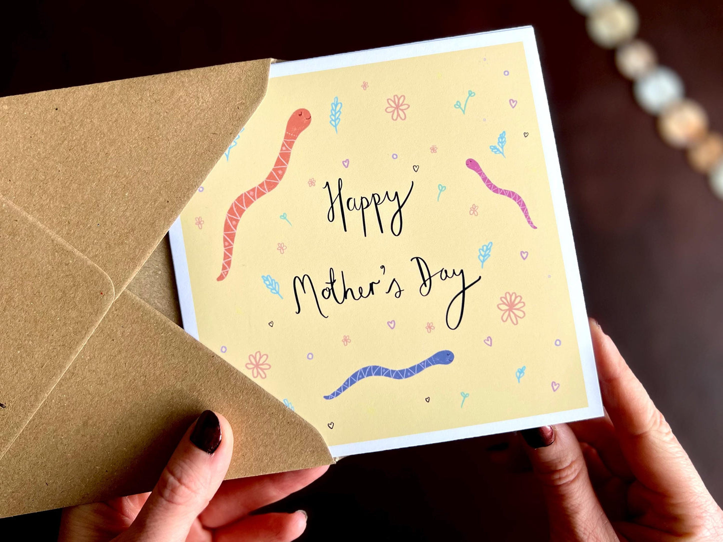 Hands holding an artisan Mother’s Day card being removed from a kraft envelope, featuring playful cartoon snakes on a lemon yellow background with handwritten style white text reading 'Happy Mother’s Day'. This version shows the alternative colour option to the navy blue design.