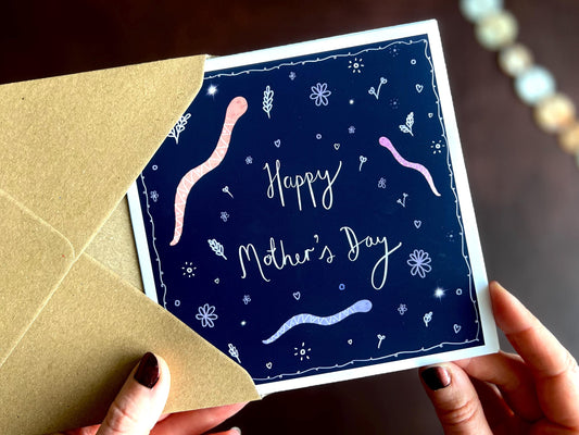 Hands holding an artisan unusual Mother’s Day card being removed from a kraft envelope, featuring playful cartoon snakes on a dark navy blue background with handwritten style white text reading 'Happy Mother’s Day'. The design has a playful dark academia inspired aesthetic.