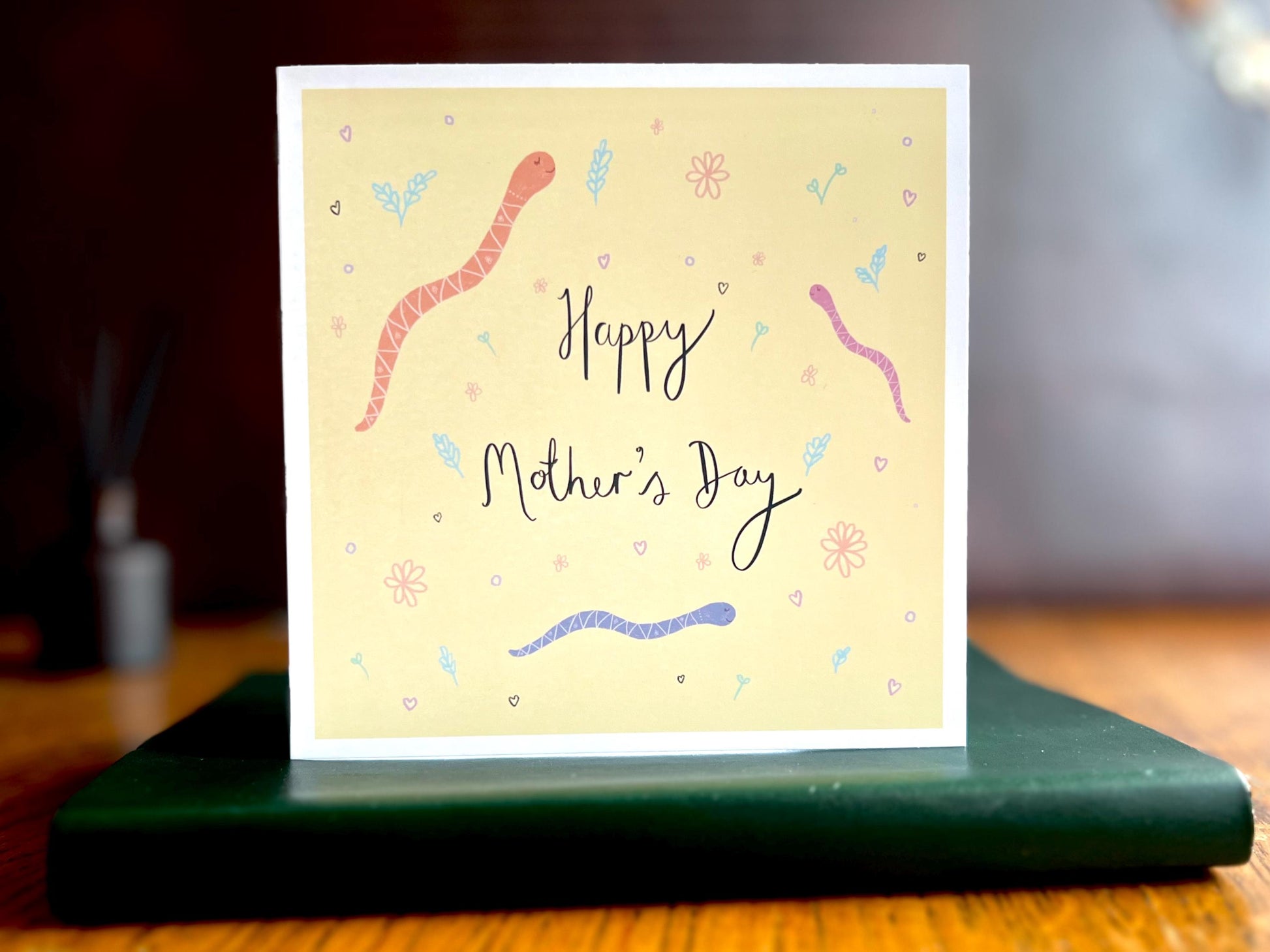 Artisan Mother’s Day card featuring playful cartoon snakes on a lemon yellow background with handwritten style white text reading 'Happy Mother’s Day', shown standing upright on a green leatherette book in a styled lifestyle display.