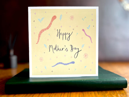 Artisan Mother’s Day card featuring playful cartoon snakes on a lemon yellow background with handwritten style white text reading 'Happy Mother’s Day', shown standing upright on a green leatherette book in a styled lifestyle display.