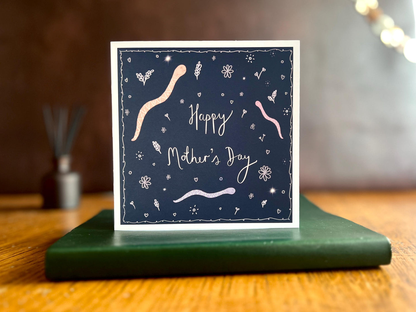 Artisan Mother’s Day card featuring playful cartoon snakes on a dark navy blue background with handwritten style white text reading 'Happy Mother’s Day', shown standing upright on a green leatherette book in a styled lifestyle display.
