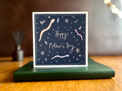 Artisan Mother’s Day card featuring playful cartoon snakes on a dark navy blue background with handwritten style white text reading 'Happy Mother’s Day', shown standing upright on a green leatherette book in a styled lifestyle display.