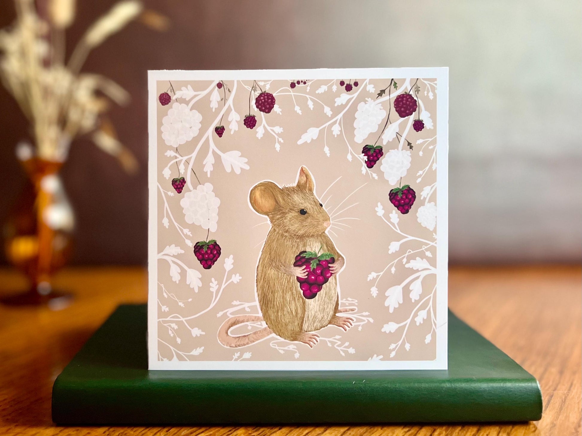 Artisan greeting card featuring a mouse holding a blackberry surrounded by a white bramble bush pattern with colourful blackberries on a pale pink background, displayed standing on a green leatherette book in a styled lifestyle setting.