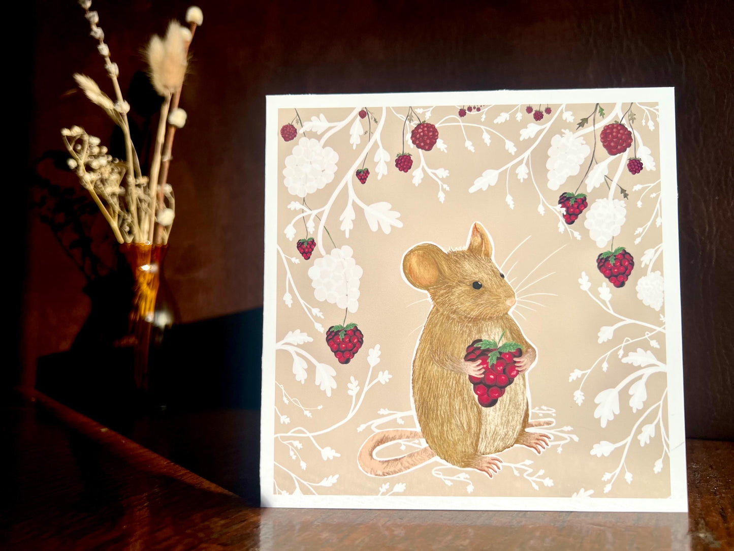 Artisan greeting card featuring a mouse holding a blackberry surrounded by a white bramble bush pattern with colourful blackberries on a pale pink background, shown in a dramatic lifestyle setup with a dark shadowy background and sunlight highlighting the card.