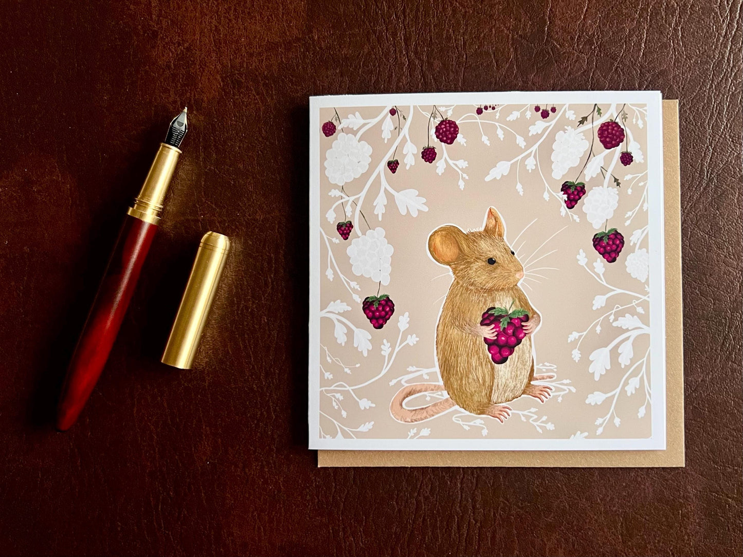 Flat lay image of an artisan greeting card featuring a mouse holding a blackberry surrounded by a white bramble bush pattern with colourful blackberries on a pale pink background, shown laid flat on a tabletop with its kraft envelope and a pen in a styled composition.