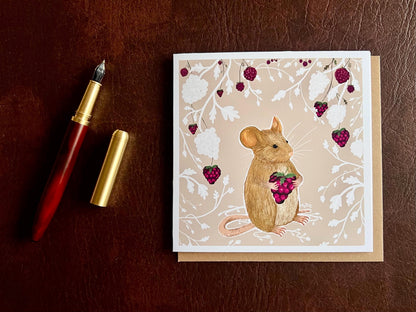 Flat lay image of an artisan greeting card featuring a mouse holding a blackberry surrounded by a white bramble bush pattern with colourful blackberries on a pale pink background, shown laid flat on a tabletop with its kraft envelope and a pen in a styled composition.