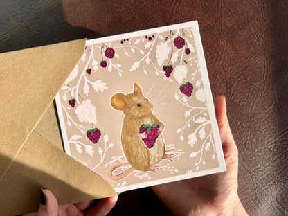 Hands holding an artisan greeting card being removed from a kraft envelope, featuring an illustration of a mouse holding a blackberry surrounded by a white bramble bush pattern with colourful blackberries forming a border on a pale pink background.