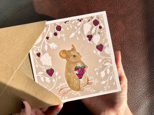 Hands holding an artisan greeting card being removed from a kraft envelope, featuring an illustration of a mouse holding a blackberry surrounded by a white bramble bush pattern with colourful blackberries forming a border on a pale pink background.